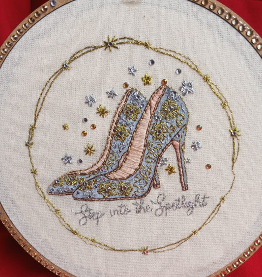Handmade embroidery of ornate high heels with gold and silver thread, star motifs, and crystal accents in a studded hoop.
